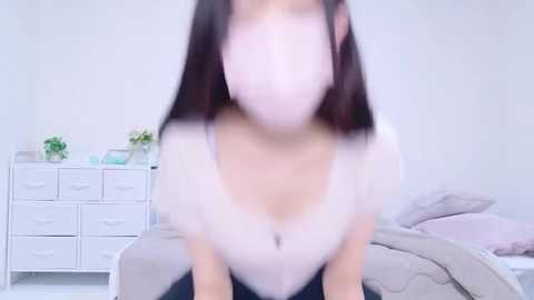 Media: A blurred video of a woman in a white top, wearing a pink face mask, sitting on a bed in a white room with a dresser and plants in the background.