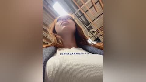 Media: Video of a young woman with long red hair, wearing a white ribbed tank top with \"FUCK YOU\" printed in black, standing in a rustic, wooden-beamed room with a bright ceiling light.