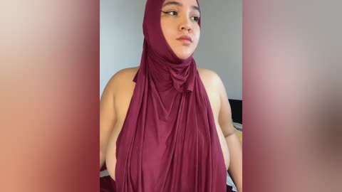 Media: Video of a young woman with medium skin tone, wearing a maroon hijab and a matching top, gazing thoughtfully to the side, with blurred background.