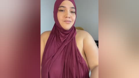 Media: Video of a smiling, light-skinned woman with medium-length hair and brown eyes wearing a maroon hijab, set against a blurred, neutral-colored background.