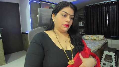 Media: Video of a South Asian woman with long black hair, fair skin, red lipstick, wearing a black top, red sari, and gold necklace, seated in a modern bedroom with a bed and window curtains.