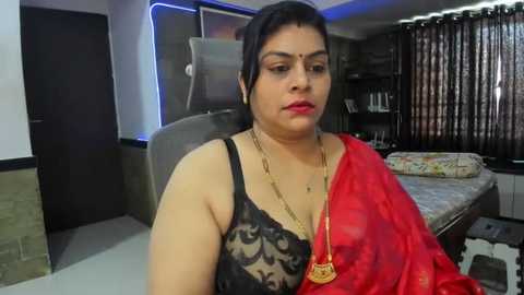 Media: Video of a middle-aged South Asian woman with short dark hair, wearing a red sari with a black lace bra, seated in a modern living room with a TV and a bed in the background.