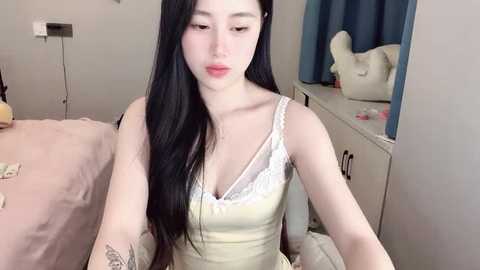 Media: A video of an East Asian woman with long black hair, wearing a beige camisole with white lace trim, sitting on a bed in a modest bedroom with light walls and a blue curtain.