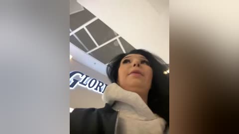 Media: A video of a woman with long black hair, wearing a black jacket and white turtleneck, captured from a low angle, with a blurred background showing part of a store sign and ceiling.