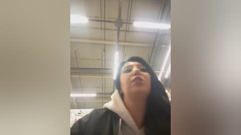 Media: A video of a young woman with long black hair, wearing a black jacket and white hoodie, indoors under fluorescent lights in a warehouse setting.