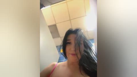 Media: Video of a young woman with long black hair, light skin, and wearing a pink bra, taken in a public restroom with blue-tiled walls and a fluorescent ceiling.