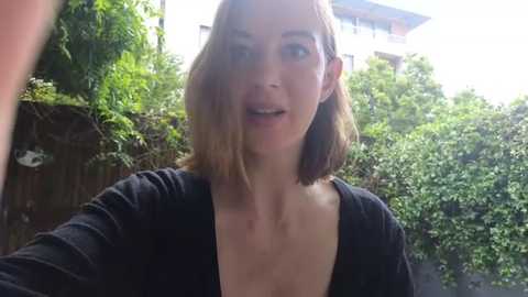 Media: A video of a Caucasian woman with light skin and shoulder-length, straight brown hair, smiling and slightly squinting, wearing a black cardigan. She is outdoors in a garden with lush green foliage and a wooden fence in the background.