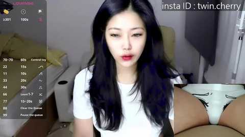 Media: A video of an Asian woman with long black hair, wearing a white top, and red lipstick, sitting on a bed, with a smartphone displaying her underwear.