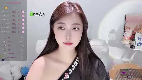 Media: Video of a young East Asian woman with straight, dark hair, fair skin, and red lipstick, wearing a black strapless dress, seated indoors with a white sofa and TV in the background.