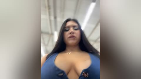 Media: A low-angle video of a curvaceous woman with long black hair and dark makeup, wearing a revealing, blue, plunging neckline top, in an industrial setting with exposed beams.