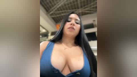 Media: Video of a curvy Latina woman with long black hair, wearing a low-cut blue tank top, revealing ample cleavage, in a modern indoor setting with exposed beams.