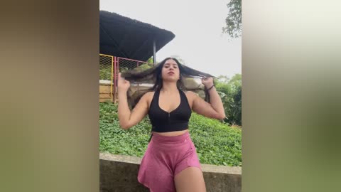 Media: Video of a young woman with long dark hair, wearing a black sports bra and pink shorts, standing outdoors in a lush garden with a chain-link fence and wooden structure in the background.