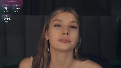 Media: Video of a young Caucasian woman with long, straight blonde hair, wearing a white top, against a dark, blurred background.