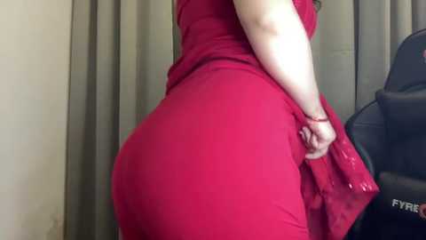 Media: Video of a plus-sized woman in a red dress, showcasing her large buttocks. She is standing in front of gray curtains and a black gaming chair.