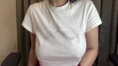 Media: Video of a woman wearing a white T-shirt with a grayscale Eiffel Tower design. Her skin is light, and she has long, dark hair. She stands against a beige curtain.