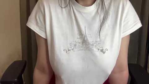 Media: Video of a woman wearing a white T-shirt with a black graphic of the Eiffel Tower. The shirt is slightly wet, revealing her breasts and nipples. Background shows beige curtains and a dark chair.