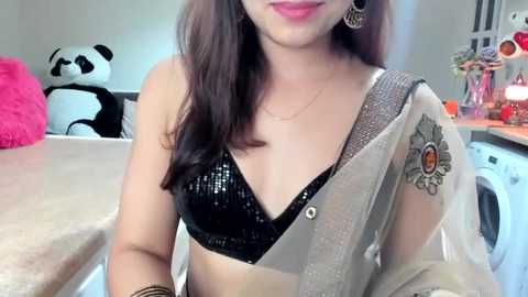 Media: Video of a smiling South Asian woman with long brown hair, wearing a black sequin bra and a beige dupatta with a floral design, seated at a kitchen counter with a panda pillow and a pink cushion in the background.