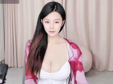 Media: A video of an East Asian woman with long, straight black hair, fair skin, and a slender build. She wears a low-cut white top revealing ample cleavage, over a pink and white floral cardigan. She sits in a beige room with curtains and a spherical lamp.