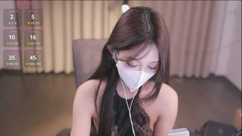 Media: Video of a young Asian woman with long black hair, wearing a white face mask, listening to earbuds, seated indoors with beige curtains and a digital clock on the left.