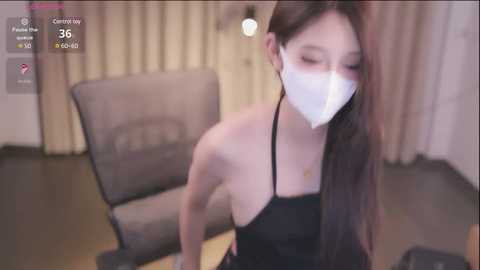 Media: A blurry photo of an Asian woman with long hair, wearing a black dress and white face mask, sitting in a beige chair in a dimly-lit room.