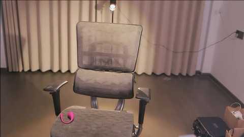 Media: Video of a medical chair with a gray cushion, lit from above, in a room with beige curtains, dark floor, and a pink vibrator on a table.