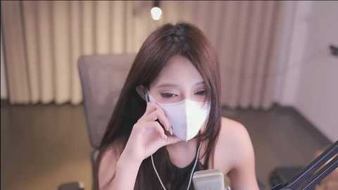 Media: Video of an East Asian woman with long brown hair, wearing a white face mask, listening to earphones, seated indoors with beige curtains and a lamp in the background.