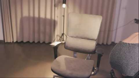 Media: Video of a modern office chair with beige upholstery and black metal frame in a dimly lit room with beige curtains and a standing lamp.