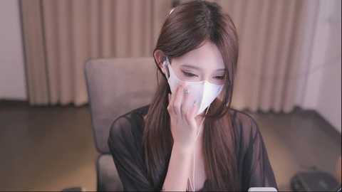 Media: Video of an Asian woman with long brown hair wearing a white surgical mask, seated in a beige armchair, indoors with beige curtains in the background.