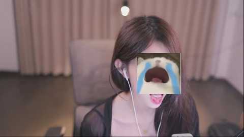 Media: Video of a young Asian woman with long brown hair, wearing a black cardigan, listening to headphones with a blue tear emoji covering her mouth, sitting on a beige chair in a dimly lit room.