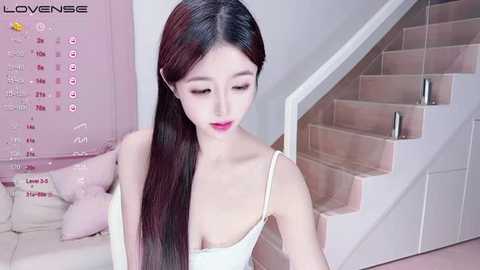 Media: A video of a young East Asian woman with long, straight, black hair, wearing a white tank top, standing on a staircase in a minimalist, pink-themed room.