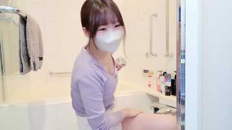 Media: Video of an Asian woman in a white bathroom, wearing a lavender sweater, white mask, and shorts, sitting on a bathtub ledge, holding a toothbrush. Background includes a towel rack, toiletries, and a bathtub.