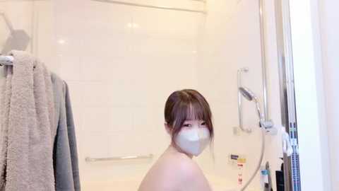 Media: A video of an Asian woman in a white-tiled bathroom, wearing a white face mask, with shoulder-length brown hair, looking back while taking a shower.