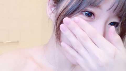 Media: Video of a young Asian woman with light skin and straight, brown hair, covering her mouth with her hand, looking slightly to the side, in a bathroom with beige tiles and a shower door.