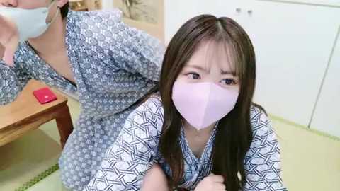 Media: A video shows an Asian woman with long brown hair wearing a blue patterned kimono, a pink mask, and a blue surgical mask, looking worried. Background features a wooden bench and white cabinets.