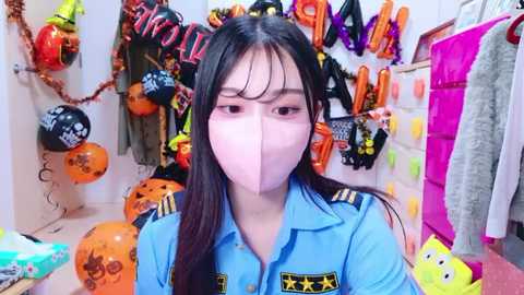 Media: Video of an East Asian woman with long black hair, wearing a blue pilot uniform and a pink face mask, surrounded by Halloween decorations, including jack-o'-lanterns, pumpkins, and festive banners.