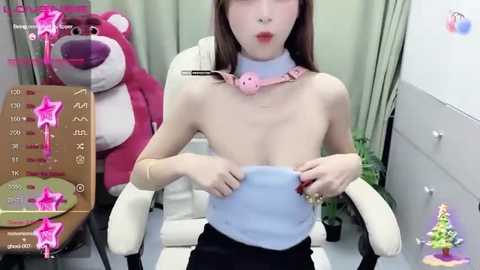 Media: A video of a young East Asian woman with fair skin, brown hair, and a slender build, wearing a light blue strapless top and black pants, seated in a white chair. Background includes a pink teddy bear and a green plant.