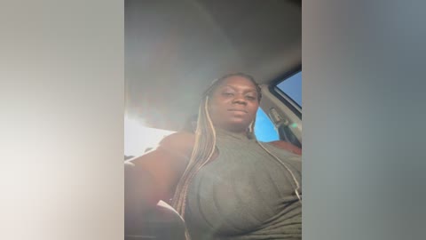 Media: Video of a Black woman with long, blonde braids, wearing a sleeveless, olive-green tank top, seated in a car under bright sunlight, her expression neutral.