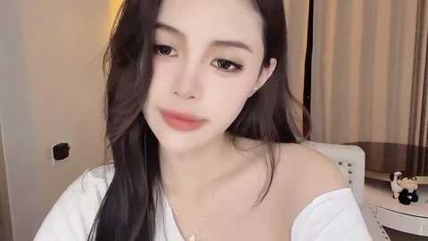 Media: Video of a young Asian woman with fair skin, long black hair, and striking dark eyes. She wears a white off-shoulder top, and the background features a beige curtain and a white cushioned chair.