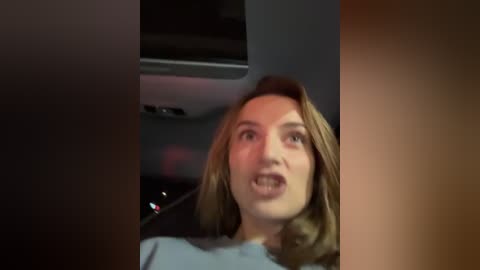 Media: A blurry video of a woman with shoulder-length brown hair, wearing a light blue shirt, inside a car, mouth open, possibly laughing.