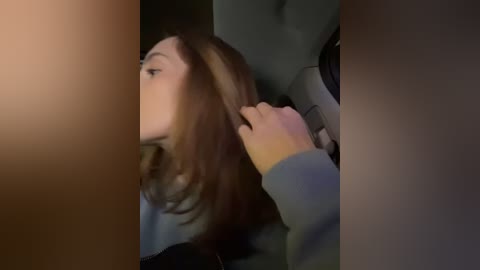 Media: Video of a woman with shoulder-length brown hair, wearing a gray sweater, seated in a car. Her hand is gently touching her hair. Background is dimly lit, focusing on her profile.