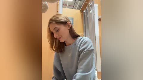 Media: Video of a young woman with light skin and shoulder-length brown hair, wearing a light gray sweatshirt, standing in a changing room with beige and orange walls, a white curtain, and a metallic ceiling.