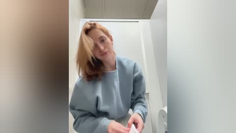 Media: Video of a fair-skinned woman with shoulder-length, light brown hair, wearing a light blue scrub top, standing in a bright, sterile bathroom with white walls and a closed door.