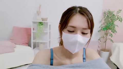 Media: Video of an Asian woman with long brown hair and light skin, wearing a blue spaghetti strap top and white face mask, seated in a softly lit, minimalist room with white walls and pastel-colored pillows.