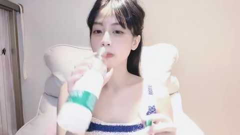 Media: Video of a young East Asian woman with straight black hair, wearing a blue and white off-shoulder top, blowing bubbles. She sits on a white chair with a white pillow. Background shows a beige wall and a wooden door.