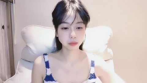 Media: Video of an Asian woman with long, straight black hair, wearing a blue and white polka-dot camisole, lying on a white pillow against a cream-colored wall.