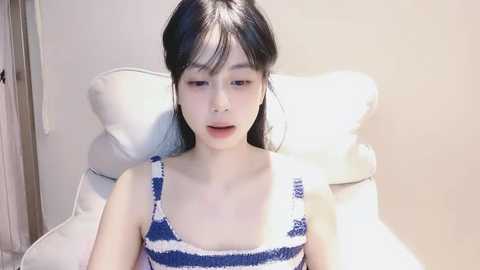 Media: Video of a young Asian woman with long black hair, wearing a blue and white striped tank top, sitting on a white chair against a plain beige wall.