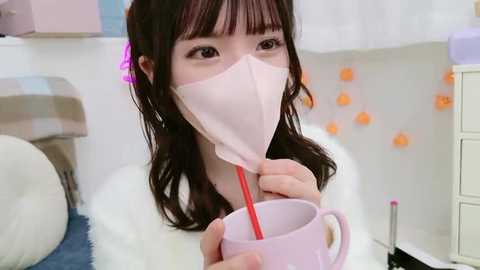 Media: A video of an Asian woman with long, dark hair in a white mask, drinking from a pink cup, in a pastel-colored room with a white bed and orange paper lanterns.