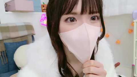 Media: Video of an Asian woman with long brown hair and bangs, wearing a white fur coat and a light pink face mask, indoors with a plaid sofa and Halloween decorations in the background.