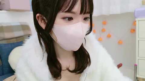 Media: Video of a young Asian woman with long black hair, wearing a pink surgical mask and white fur-lined coat, standing indoors near a white wall adorned with orange lights.