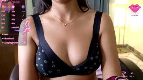 Media: Video of a woman with medium skin tone, wearing a black bra with white floral patterns, indoors with a lit lamp and partially visible screen displaying social media statistics.